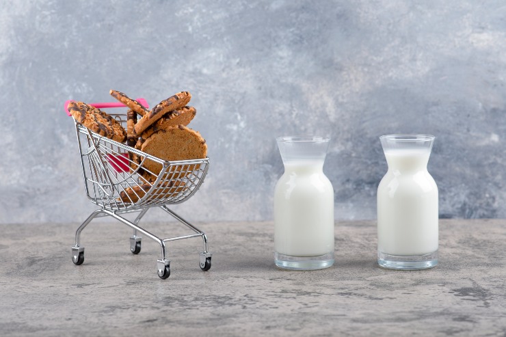 Due bottiglie di latte in vetro accanto a un piccolo carrello della spesa con biscotti al cioccolato su un tavolo